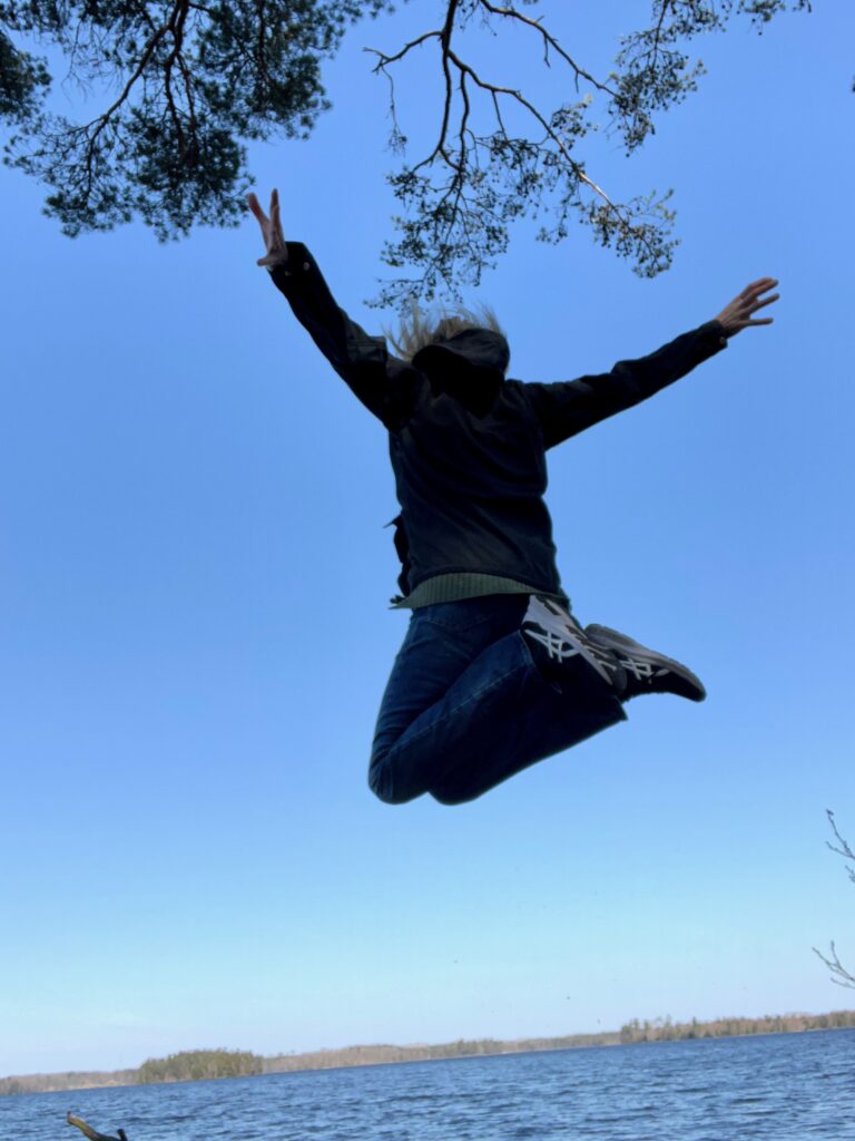 Person hoppar upp i luften mot en blå himmel vid vattnet.